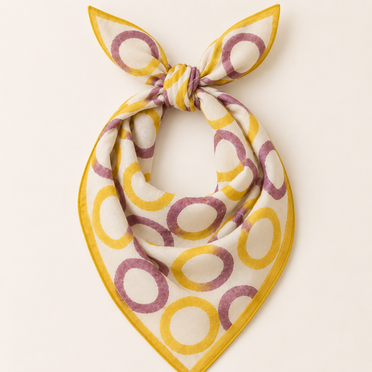 A square scarf tied in a knot, featuring a pattern of yellow and dusty purple watercolor-style circles on a warm cream background, set against a solid off-white backdrop.