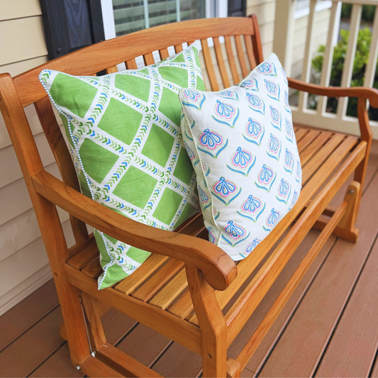 Two decorative throw pillows on a wooden porch bench. One pillow is green with a white geometric lattice pattern and blue accents, and the other is white with a blue butterfly pattern.