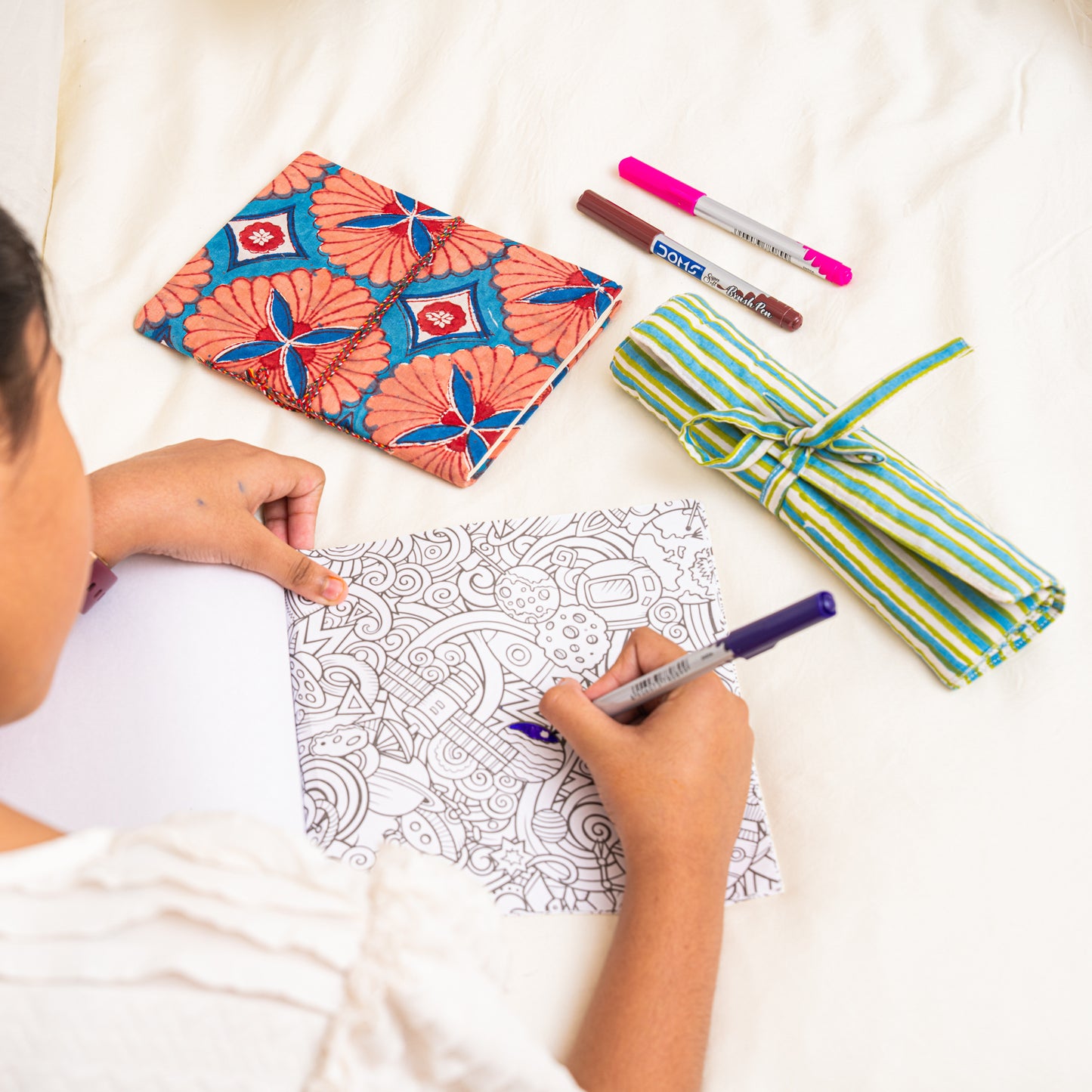 Person coloring a design with markers on a drawing surface, surrounded by cotton hand block printed journal and roll-up organizer  on a white surface 