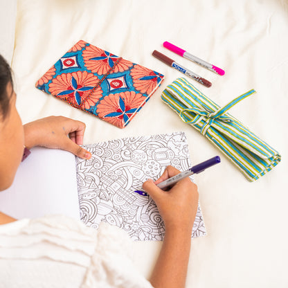 Person coloring a design with markers on a drawing surface, surrounded by cotton hand block printed journal and roll-up organizer  on a white surface 