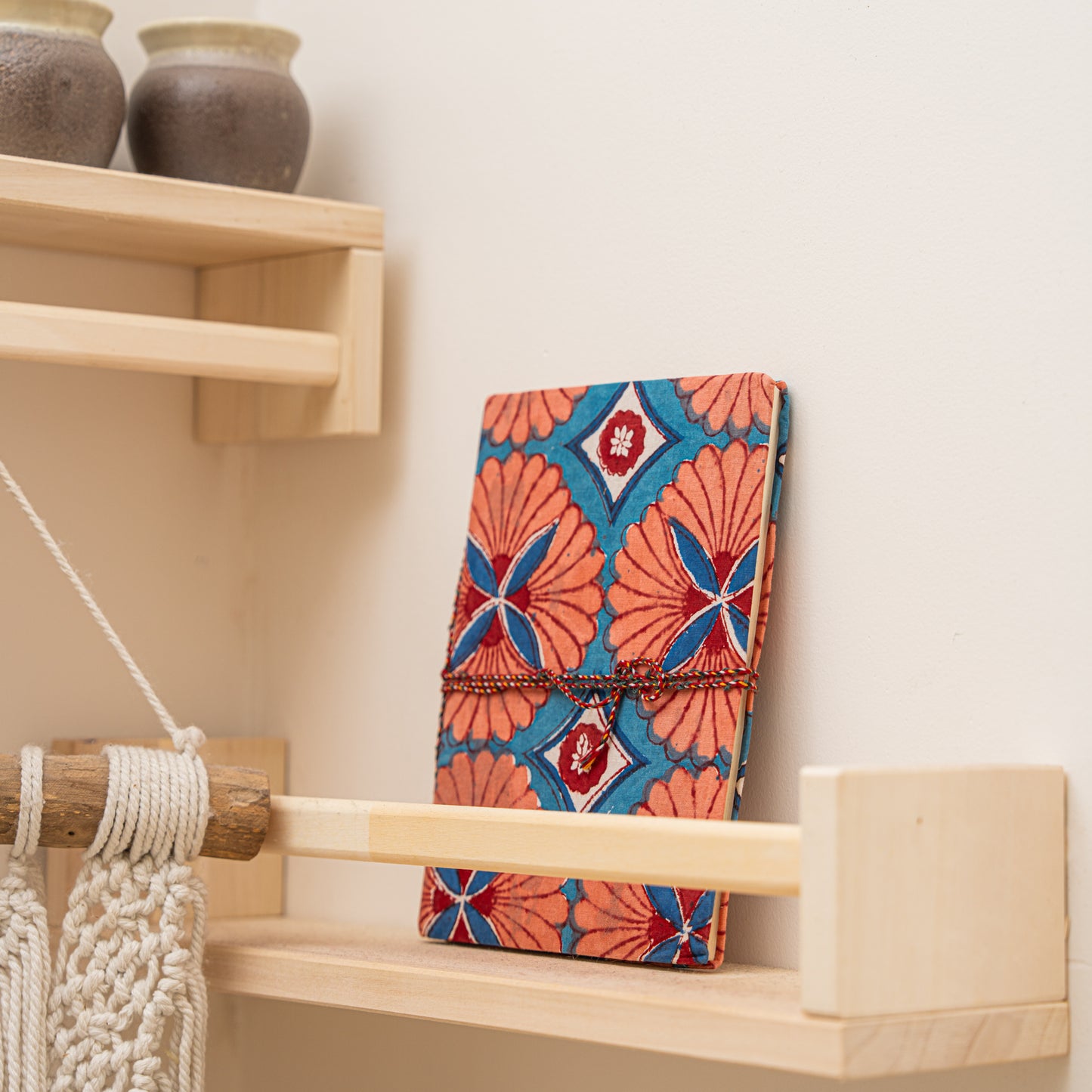 Colorful patterned journal with hand block printed pattern on cotton, placed  on a wooden shelf with ceramic jars and macrame wall hanging.