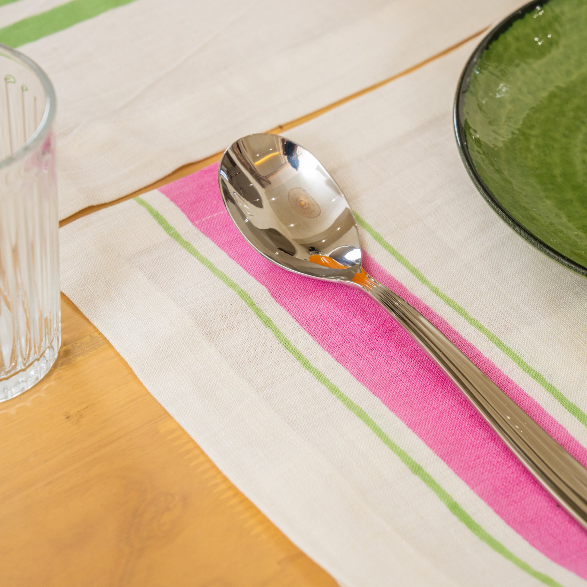 Silver spoon on a pink and green striped hand block printed linen placemat with a glass and bowl in the background