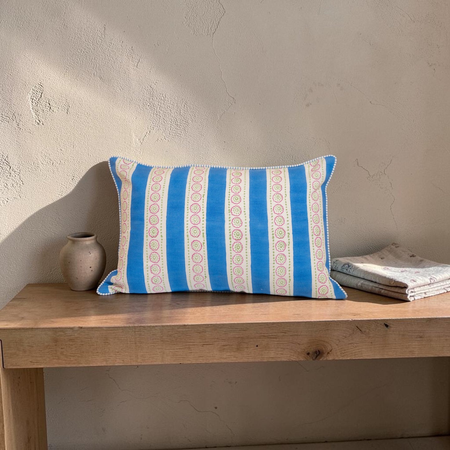 Hand block printed linen pillow cover with blue stripes and floral motifs in natural dyes placed on  a wooden bench with a neutral wall background