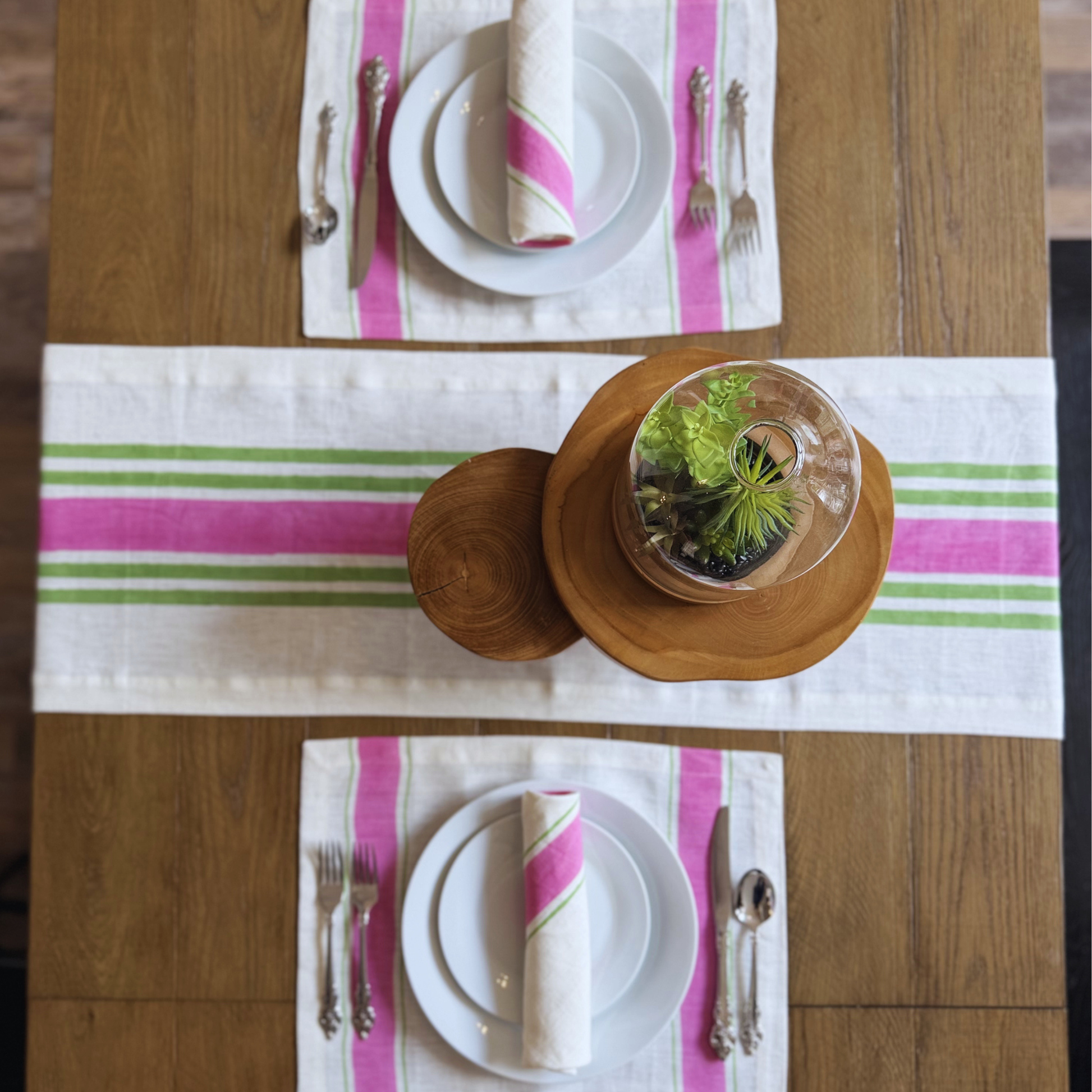 Table setting with pink and green striped hand block printed linen placemats, plates, and cutlery on a wooden table.