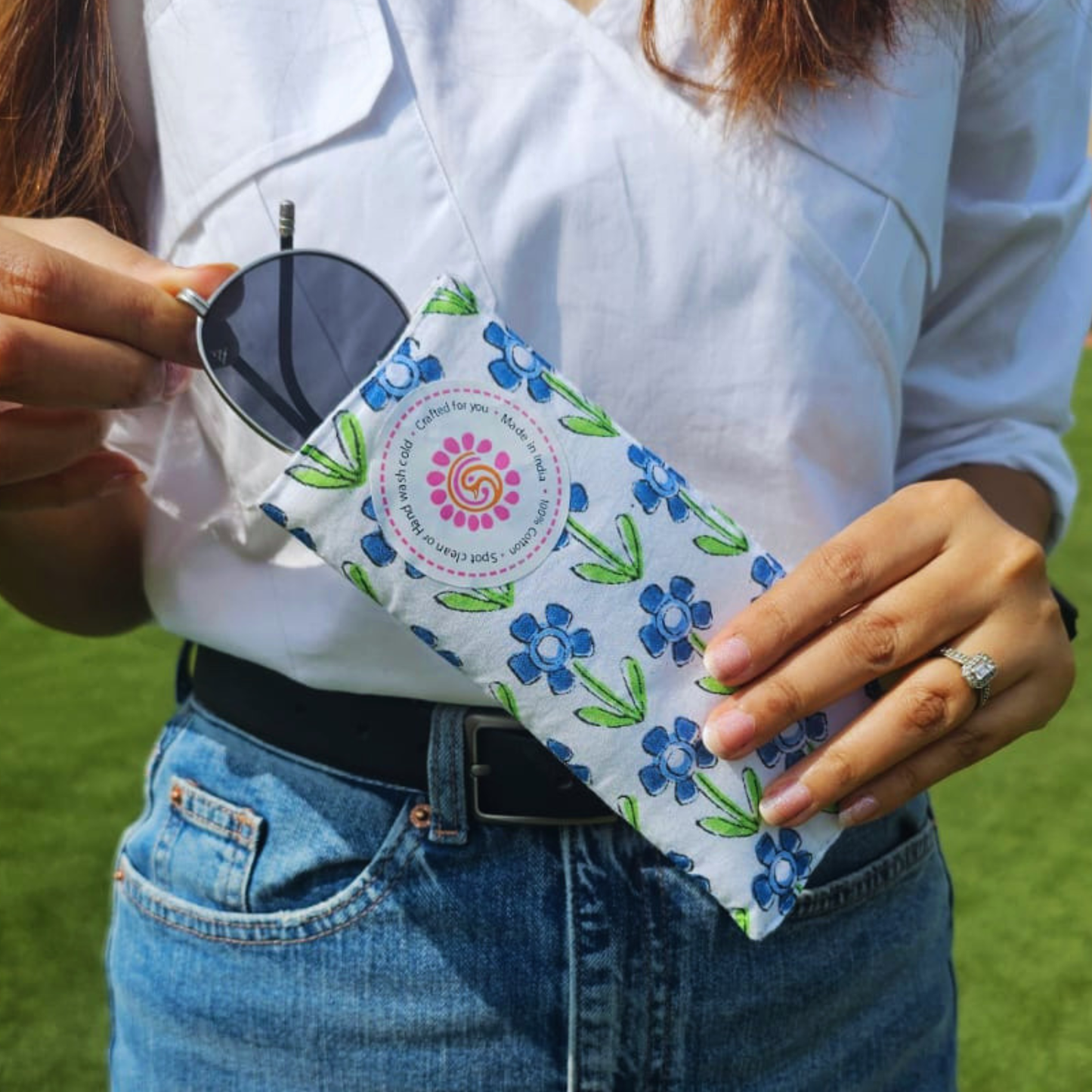 Person holding a floral-patterned hand block printed pouch with sunglasses, wearing a white shirt and blue jeans outdoors.