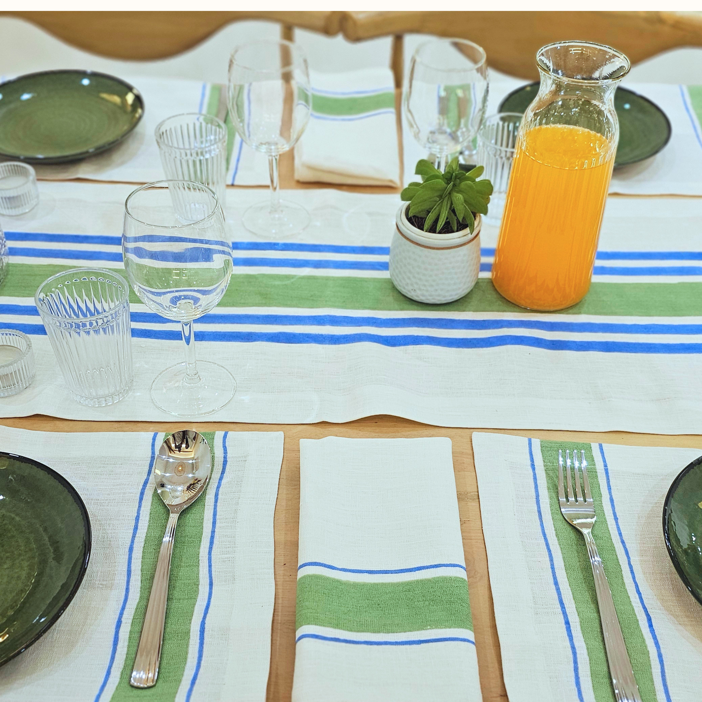 Dining table set with a blue and green hand block printed linen striped tablecloth, glasses, plates, and a carafe of orange juice.